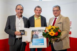 3 Männer stehen nebeneinander, zwei halten Schriftstücke und einer einen Blumenstrauß in der Hand.