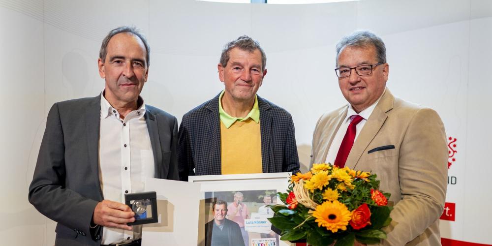 3 Männer stehen nebeneinander, zwei halten Schriftstücke und einer einen Blumenstrauß in der Hand.